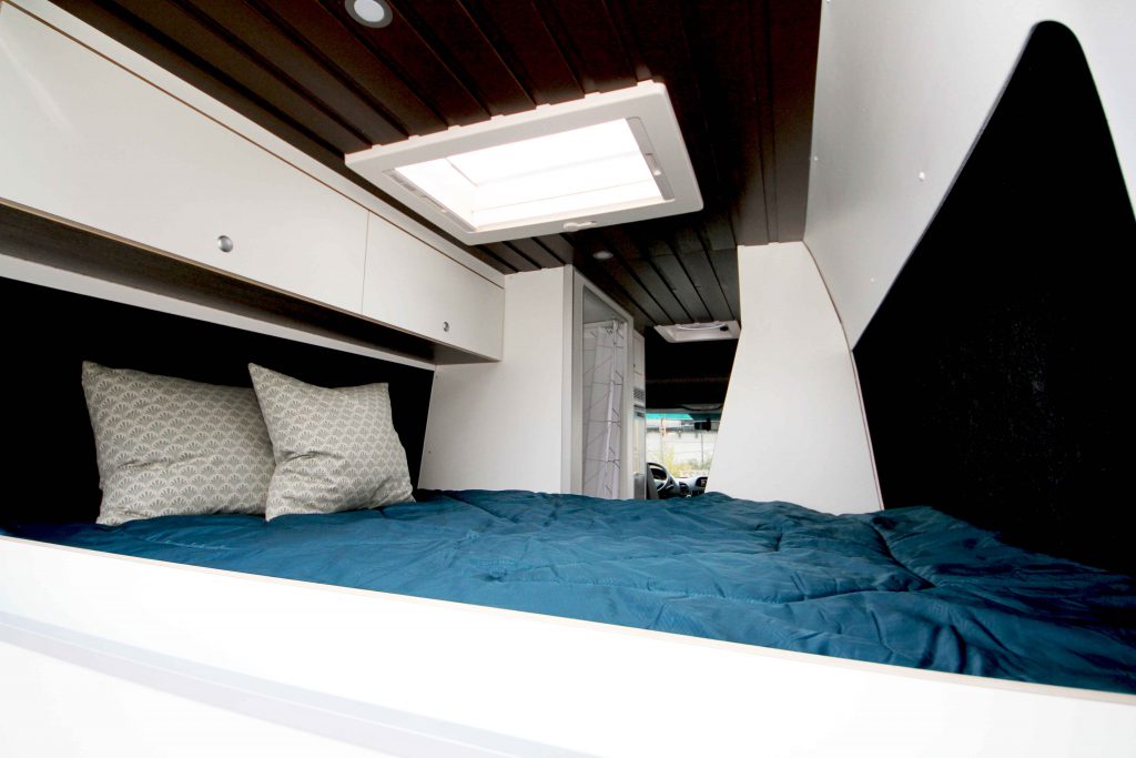 Der Mercedes Sprinter von Innen mit Blick aufs Bett, mit Leichtbau Sperrholz weiß beschichtet, brauner Deckenverkleidung für eine gemütliche Atmosphäre und Dachfenster für ein lichtdurchflutetes, modernes Raumgefühl.