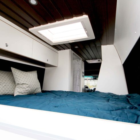 Der Mercedes Sprinter von Innen mit Blick aufs Bett, mit Leichtbau Sperrholz weiß beschichtet, brauner Deckenverkleidung für eine gemütliche Atmosphäre und Dachfenster für ein lichtdurchflutetes, modernes Raumgefühl.